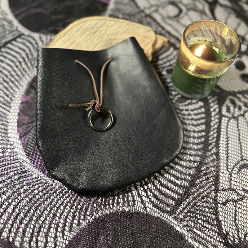 A large black leather drawstring pouch with a silver carabiner ring, resting on patterned lace beside a lit candle.