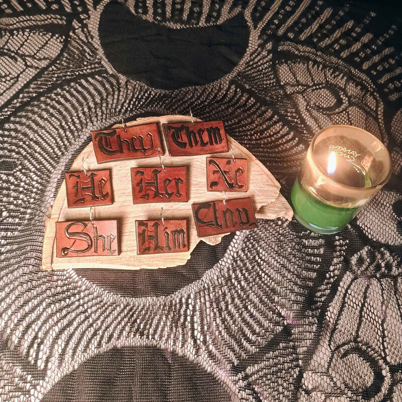 Leather pronoun earrings hand-tooled with They, Them, He, Her, Xe, Xhu, She and Him, laid on a wooden board beside a lit candle.