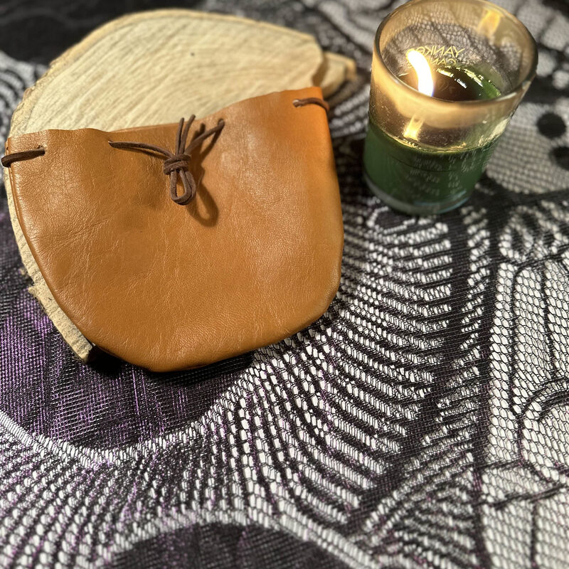 A small tan leather drawstring pouch tied with a dark leather cord, resting on a wooden slice beside a lit green candle.